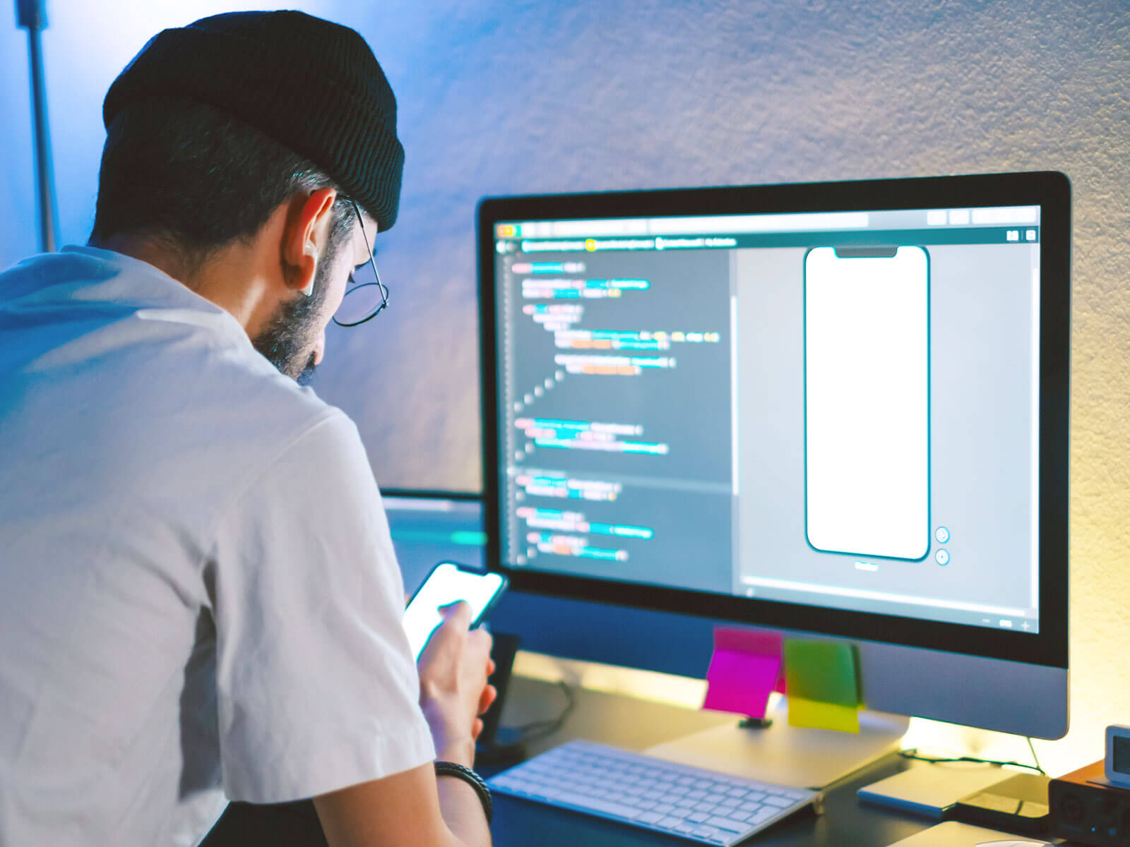 Developer working on an app, looking at mobile phone rendering and code on a computer screen