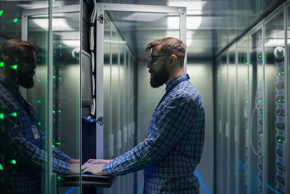 Adult bearded system administrator in glasses working on laptop and correcting errors in server hardware.