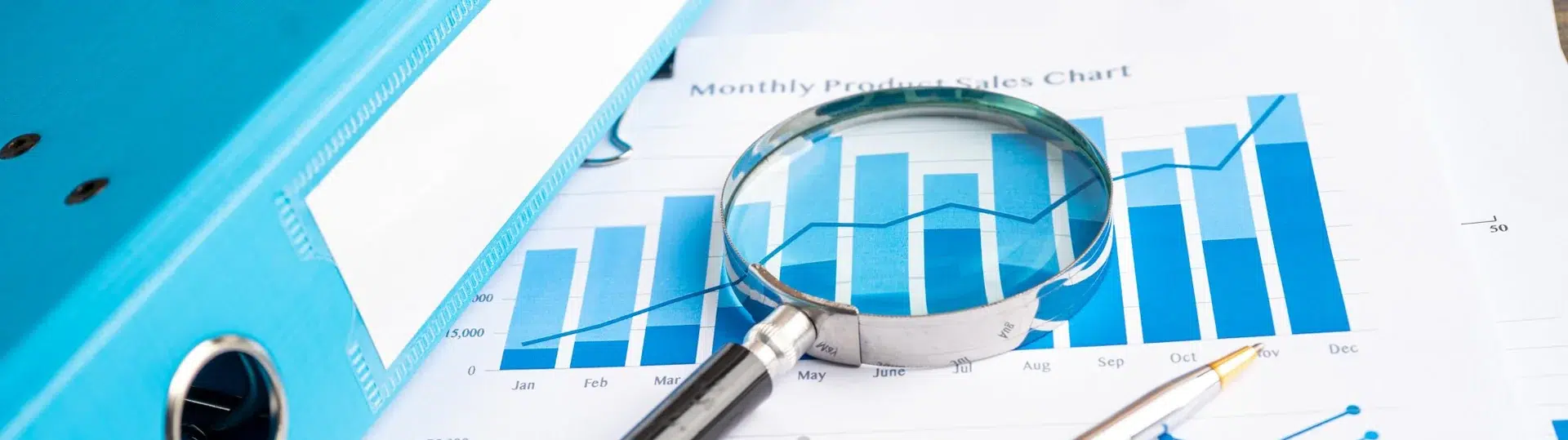 Business analyst using a magnifying glass to examine monthly product sales charts and data reports