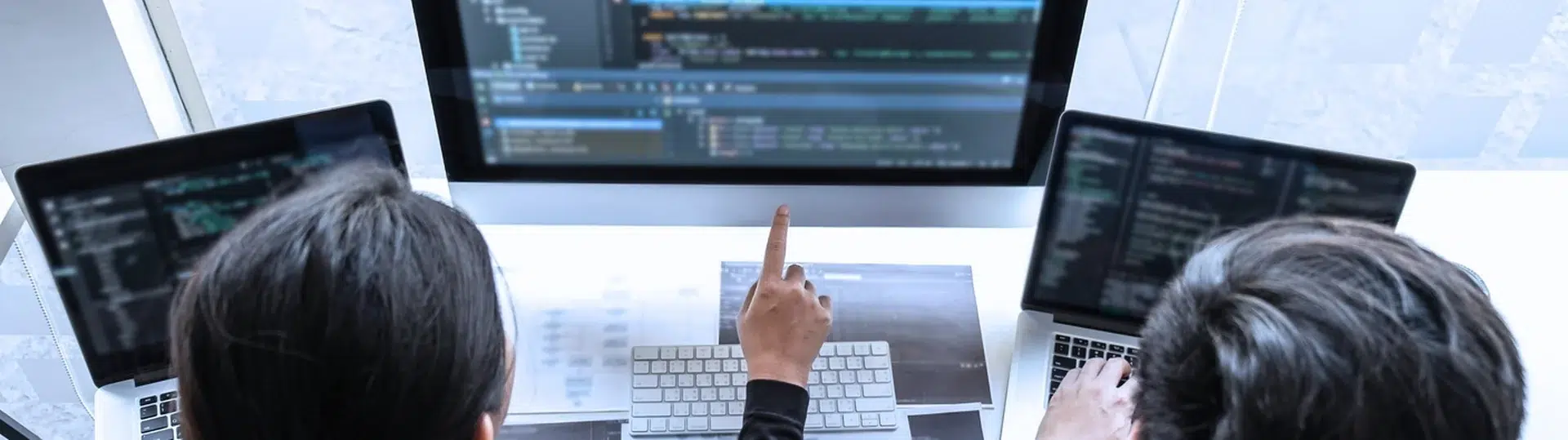 Overhead view of two engineers reviewing and pointing at code on multiple screens during collaborative session