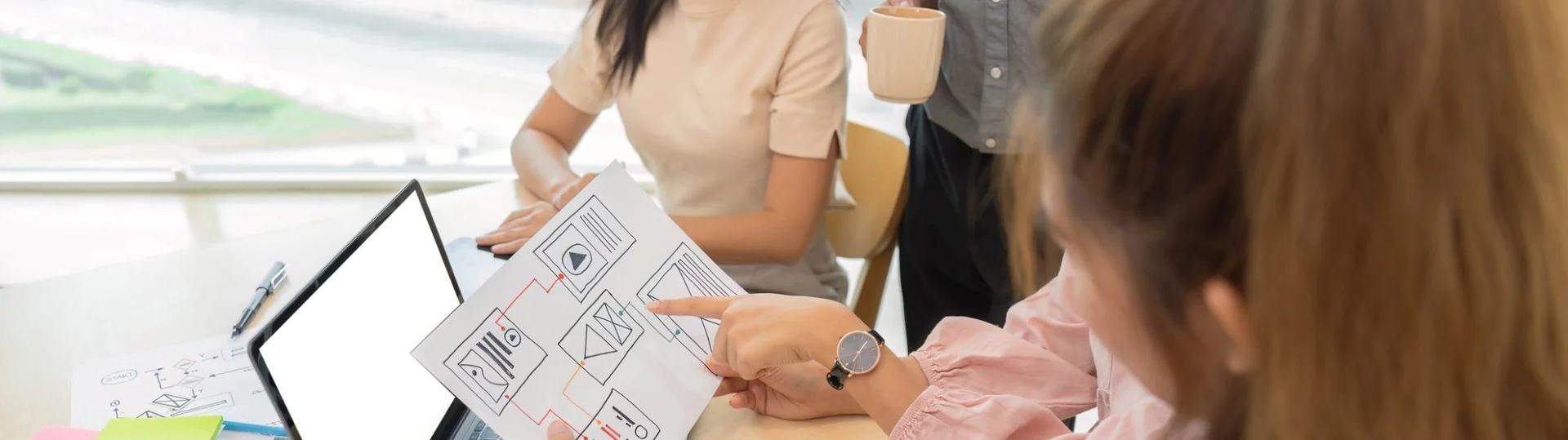 Three-person UX team collaborating on mobile app wireframe printouts at a bright office table