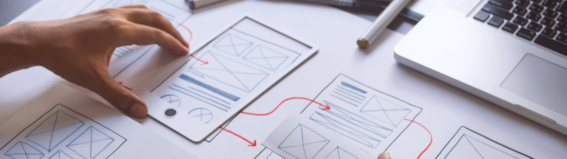 Designer arranging hand-drawn mobile app wireframe sketches with red flow arrows on a desk
