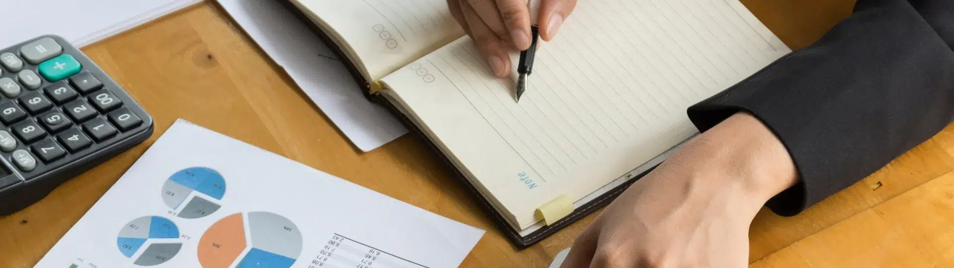 Strategic planner writing in a notebook alongside printed data charts and a laptop on a wooden desk