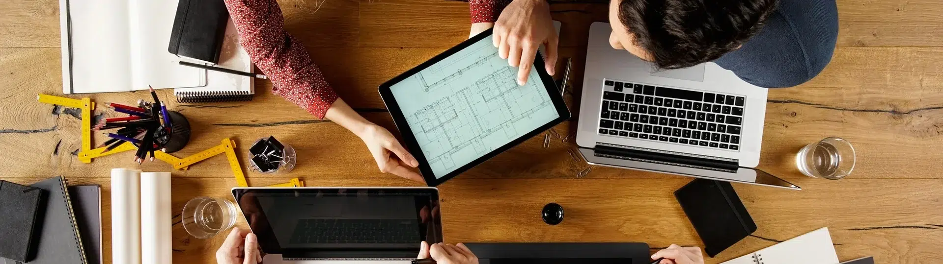 Overhead view of a cross-functional team collaborating with tablets, laptops, and blueprints on a wooden table
