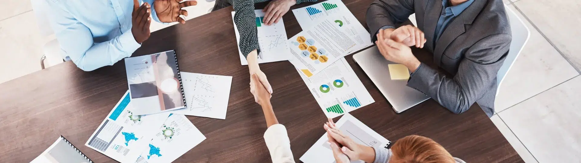 Overhead view of a team collaborating around a table with data reports, handshake in foreground