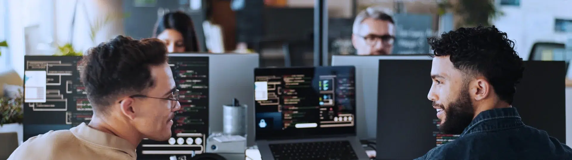 Two developers pair programming and discussing AI-assisted code solutions at workstations with multiple monitors