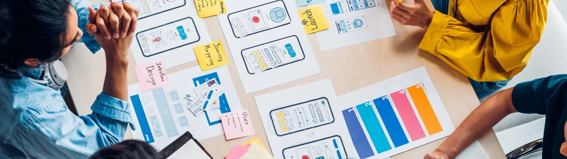 UI/UX design team collaborating over wireframe layouts, color palettes, and design materials spread across a table