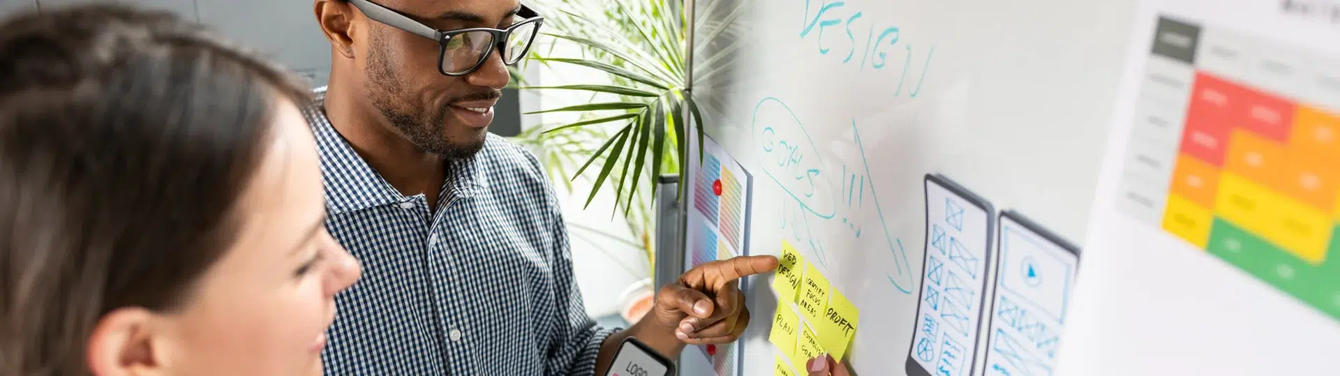 Two UX designers reviewing mobile wireframes and sticky notes on a whiteboard during a design planning session